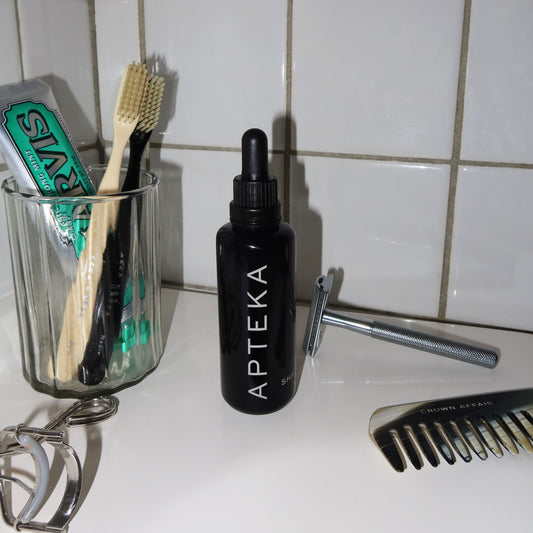 A black dropper bottle labeled APTEKA sits on a bathroom counter with a glass cup holding toothbrushes, a razor, an eyelash curler, and a comb, against a white tiled wall.