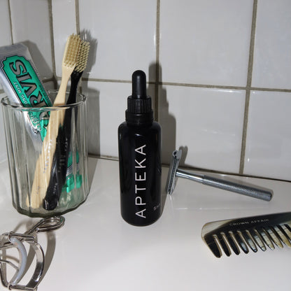 A black dropper bottle labeled APTEKA sits on a bathroom counter with a glass cup holding toothbrushes, a razor, an eyelash curler, and a comb, against a white tiled wall.