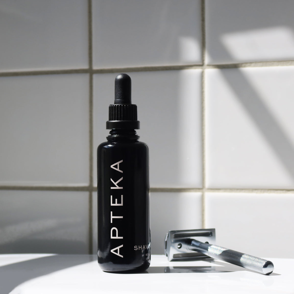 A black dropper bottle labeled APTEKA stands on a white surface against a white tiled wall, with a silver safety razor resting beside it in soft sunlight and shadow.