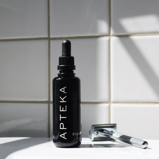 A black dropper bottle labeled APTEKA stands on a white surface against a white tiled wall, with a silver safety razor resting beside it in soft sunlight and shadow.